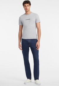 Sivý tričko s krátkym rukávom s logom "GUESS JEANS", kombinované s tmavomodrými džínsami a bielymi teniskami, stojace na jednoduchom pozadí.