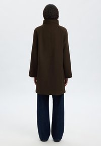 Manteau marron avec un col montant et de larges manches, fabriqué dans un tissu doux. Porté sur un jean bleu foncé ample, vu de dos.
