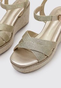Sandali con zeppa in finitura oro metallizzato, caratterizzati da incroci in tessuto intrecciato e una suola a piattaforma simile al juta. Sottopiede liscio e lucido.