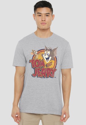 Man wearing light gray T-shirt with colorful Tom and Jerry cartoon graphic and beige pants, standing against a plain background.