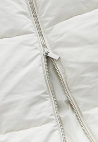 Veste matelassée blanche, avec un détail de fermeture éclair argentée. Texture lisse et brillante, avec des sections quiltées et un design léger.