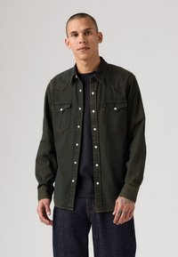 Camicia di denim verde scuro con bottoni a pressione, due tasche sul petto e cuciture a contrasto. Indossata sopra una t-shirt nera e jeans blu.