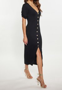 Femme portant une robe midi noire à boutons dorés sur le devant et une fente à l'avant, associée à des sandales à talons hauts avec lanières dorées.