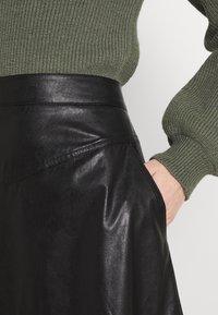 Musta nahkahame, jossa on sileä pinta, korkea vyötärö ja vinot taskut, yhdistetty vihreään ribbipaita.
