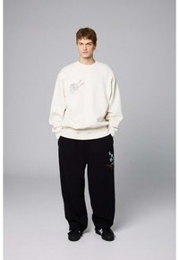 Kremowy sweatshirt z grafiką pałeczek na klatce piersiowej, zestawiony z czarnymi spodniami z kolorowymi haftami. Luźny krój, wygodny materiał.