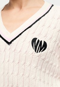 Κλείσιμο πλάνου ενός κρεμ πουλόβερ V-λαιμόκοψη με πλέξη καλωδίου, με μαύρες και ροζ ρίγες στο γιακά και ένα μαύρο λογότυπο σε σχήμα καρδιάς στο στήθος.