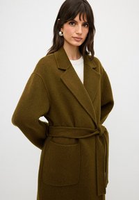 Olivgrön ullkappa med bred krage, bälte och två framfickor. Har en mjuk textur och lös passform.