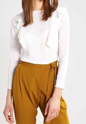 Femme portant un haut blanc à manches longues avec des volants et un pantalon jaune moutarde taille haute noué à la taille.