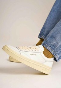 Sneakers in pelle bianca con accenti beige, caratterizzati da un design con lacci e logo del marchio in rilievo. Indossati con jeans blu e calze testurizzate.
