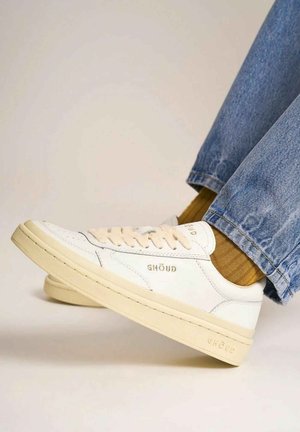 Sneakers in pelle bianca con accenti beige, caratterizzati da un design con lacci e logo del marchio in rilievo. Indossati con jeans blu e calze testurizzate.
