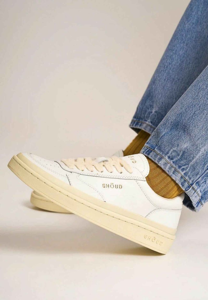 Sneakers in pelle bianca con accenti beige, caratterizzati da un design con lacci e logo del marchio in rilievo. Indossati con jeans blu e calze testurizzate.