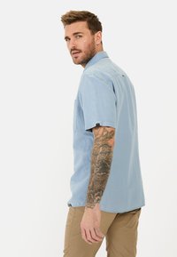 Lichtblauw kort mouw overhemd met een klein borstzakje en een subtiele logo-detail. Draagt met tan kleurige broek.