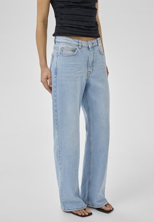 Személy világoskék bőszárú farmert, fekete fodros ujjatlan felsőt és fekete négyzet alakú orrú szandált visel, egyszínű háttér előtt áll.