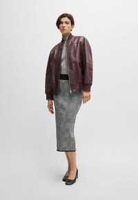 Veste bomber en cuir bordeaux avec des poignets côtelés, col roulé gris, jupe crayon à carreaux et chaussures à talons noires. Textures variées et lignes épurées.