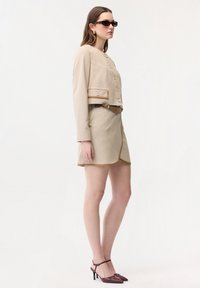 Donna con giacca beige abbottonata e gonna coordinata con cintura marrone, occhiali da sole scuri e scarpe con tacco alto color bordeaux, in piedi di profilo.