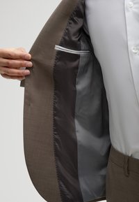Osoba trzymająca otwartą brązową marynarkę garnituru, ukazującą ciemnoszarą satynową podszewkę i wewnętrzną kieszeń, ubrana w białą koszulę i pasujące brązowe spodnie.