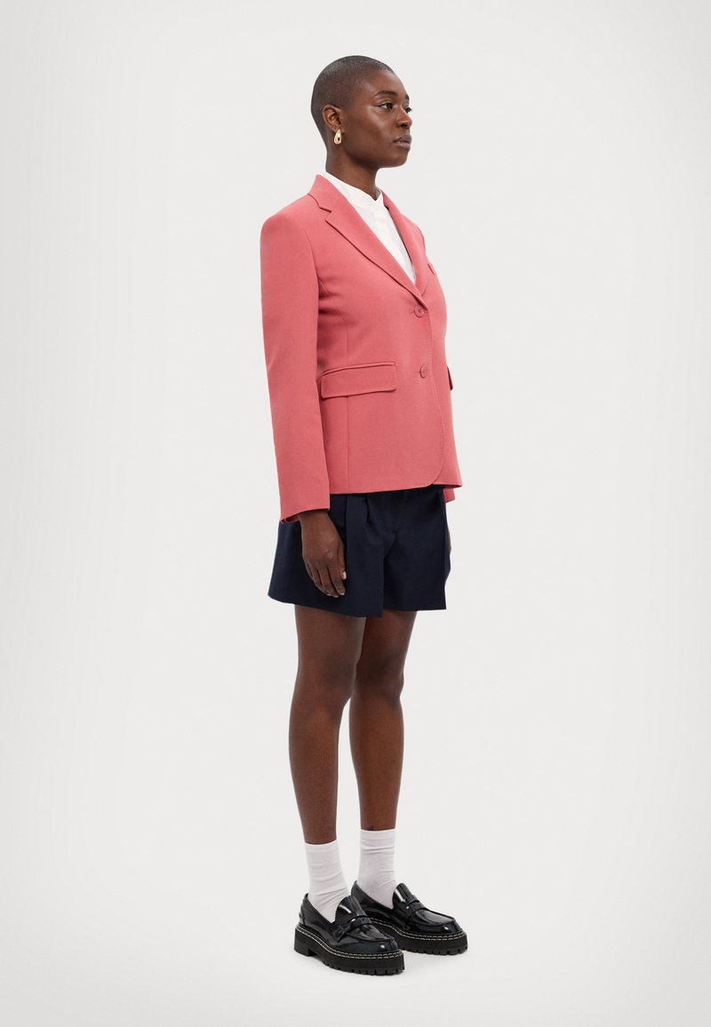 Blazer in pink fabric with structured design, featuring front pockets and a single button. Paired with navy shorts and black platform shoes.