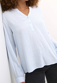 Chemise à manches longues à rayures bleu clair avec col en V, dotée d'une patte de boutonnage et d'un ourlet légèrement courbé. Tissu doux, coupe décontractée.