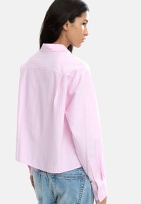 Jasnoróżowa, paskowana, oversize'owa koszula z kołnierzykiem i guzikami przy mankietach, z zaokrąglonym dołem i luźnym krojem, w komplecie z niebieskimi dżinsami.