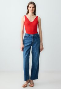Body rosso a coste con scollatura a V, abbinato a jeans blu a vita alta con gamba dritta e sandali beige su sfondo uniforme.