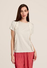 T-shirt gris clair avec un col rond, associ é à un pantalon corail à motif de feuilles. Un gilet en dentelle blanche drape le t-shirt.