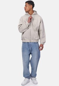 Hoodie zippé beige clair avec un logo, manches larges et une poche avant, associé à un jean bleu clair ample et des baskets blanches.