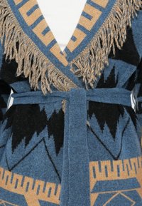 Cardigan tricotat cu model albastru și negru, cu margine geometrică bej și franjuri, legat în talie peste o bluză albă.