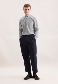 Camicia grigia chiara con bottoni e piccoli quadretti, abbinata a pantaloni blu scuro plissettati e scarpe di pelle nere, il tutto su uno sfondo neutro.