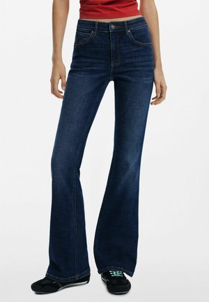 Flared dark blue denim jeans with a smooth texture, five-pocket design, and metal button closure. Paired with black footwear.
