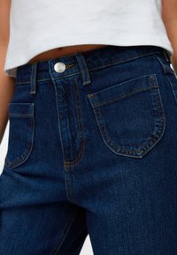 Jeans de mezclilla azul de talle alto con diseño tradicional de cinco bolsillos, con costuras naranjas y cierre de botón plateado. Textura suave.
