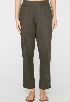 Kobieta w ciemnooliwkowych, wysokich w talii, prostych spodniach oraz płaskich sandałach z plecionymi paskami, stojąca na tle jednolitego jasnego tła.