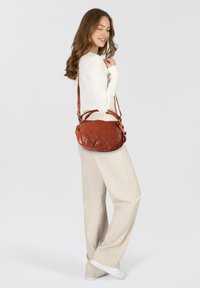 Bolso bandolera de cuero marrón con una textura suave, forma redondeada, correa ajustable y detalles decorativos en el hardware.