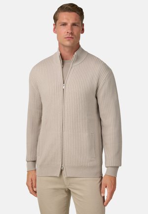 Beige gerippter Zipper-Pullover mit hohem Kragen. Verfügt über zwei Fronttaschen und strukturierten Stoff. Trägt man über einem hellen Hemd.