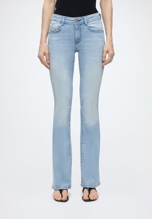 Slim fit lyseblå jeans båret med sort top og sorte åbne sandaler, vist fra taljen til fødderne på ensfarvet baggrund.