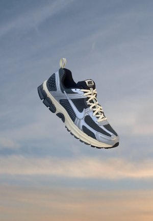 Chaussure de course Nike grise et noire avec des lacets crème flottant sur un fond de ciel nuageux au coucher du soleil.