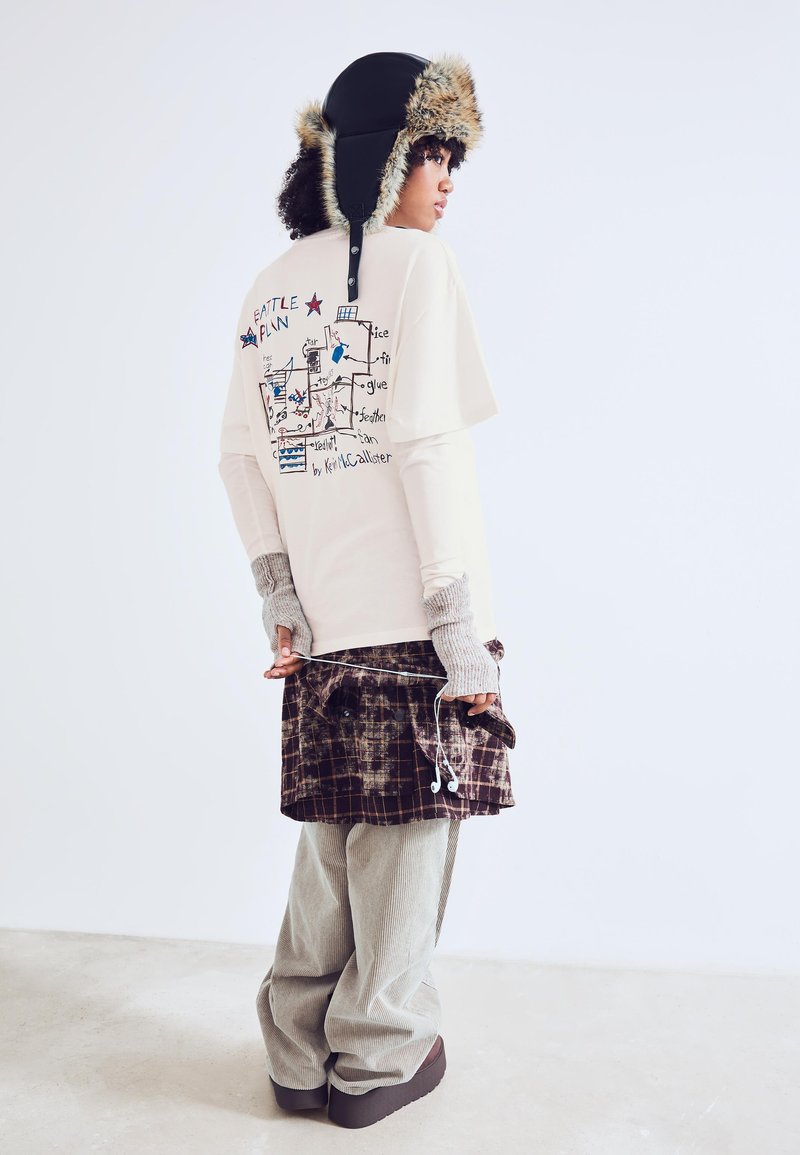 Tricou crem cu imprimeu grafic, purtat sub o fustă scurtă cu carouri maro. Pantaloni gri din catifea și o pălărie neagră cu blană la margine completează ținuta.