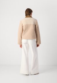 Veste en polaire douce beige et crème avec une finition texturée, dotée d'un col droit et de poignets élastiques, associée à un pantalon blanc à jambe large.