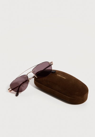 Rimless sunglasses with purple lenses resting on a brown suede Tom Ford case on a white surface.