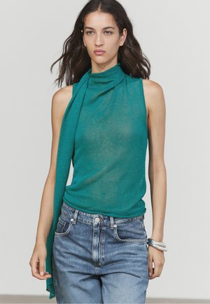 Vrouw met lang donker haar die een mouwloos turquoise top met sjaaldetail en hoogwater blauwe jeans draagt, loopt zelfverzekerd.