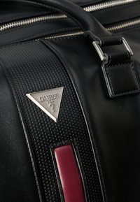 Fekete bőr táska texturált sávval, ezüst logólemezsel és piros díszítéssel. Ezüst cipzárokkal és masszív fogantyúval rendelkezik.