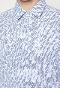 Chemise blanche à boutons avec col pointu et motif vertical à tirets bleu marine portée par une personne visible du cou jusqu’au milieu de la poitrine.