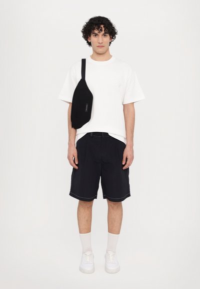 Witte t-shirt met korte mouwen en geborduurde tekst, gecombineerd met zwarte, op maat gemaakte shorts, witte sneakers en een zwarte schoudertas.
