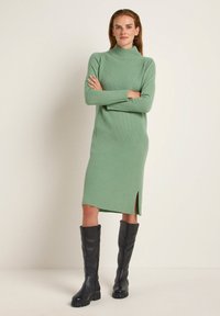 Mintgrünes geripptes Kleid mit hohem Hals, langen Ärmeln und einem seitlichen Schlitz. Kombiniert mit knielangen schwarzen Lederstiefeln.