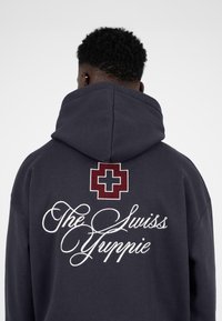 Donkerblauwe hoodie met grote geborduurde witte letters "The Swiss Yuippie" en een bordeauxrode Zwitserse kruis op de achterkant. Zachte stoftextuur.