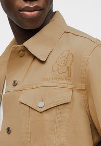 Κοντινό πλάνο ατόμου που φοράει μια μπεζ τζιν ζακέτα με κεντητό λουλούδι και τη λέξη «marimekko» στην περιοχή της τσέπης στο στήθος.