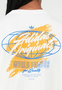 Žmogus dėvi baltą marškinį su mėlynu ir oranžiniu "Adidas Originals" logotipu, pasaulio žemėlapiu ir tekstu "Pasaulinis garsas dėl kokybės 1972" ant nugaros.