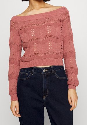 Suéter de punto rosa con escote off-the-shoulder, con patrones texturizados y detalles de punto abierto, combinado con unos pantalones vaqueros de talle alto en azul oscuro.