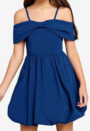 Mädchen trägt ein ärmelloses dunkelblaues Kleid mit dünnen Trägern, schulterfreiem Schleifendetail und einem gerafften, leicht gepufften Rock.
