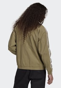 Olivgrön, lätt jacka med vita sidostråk, elastiska ärmslut och en avslappnad passform. Har en smidig textur och enkel sömnad.