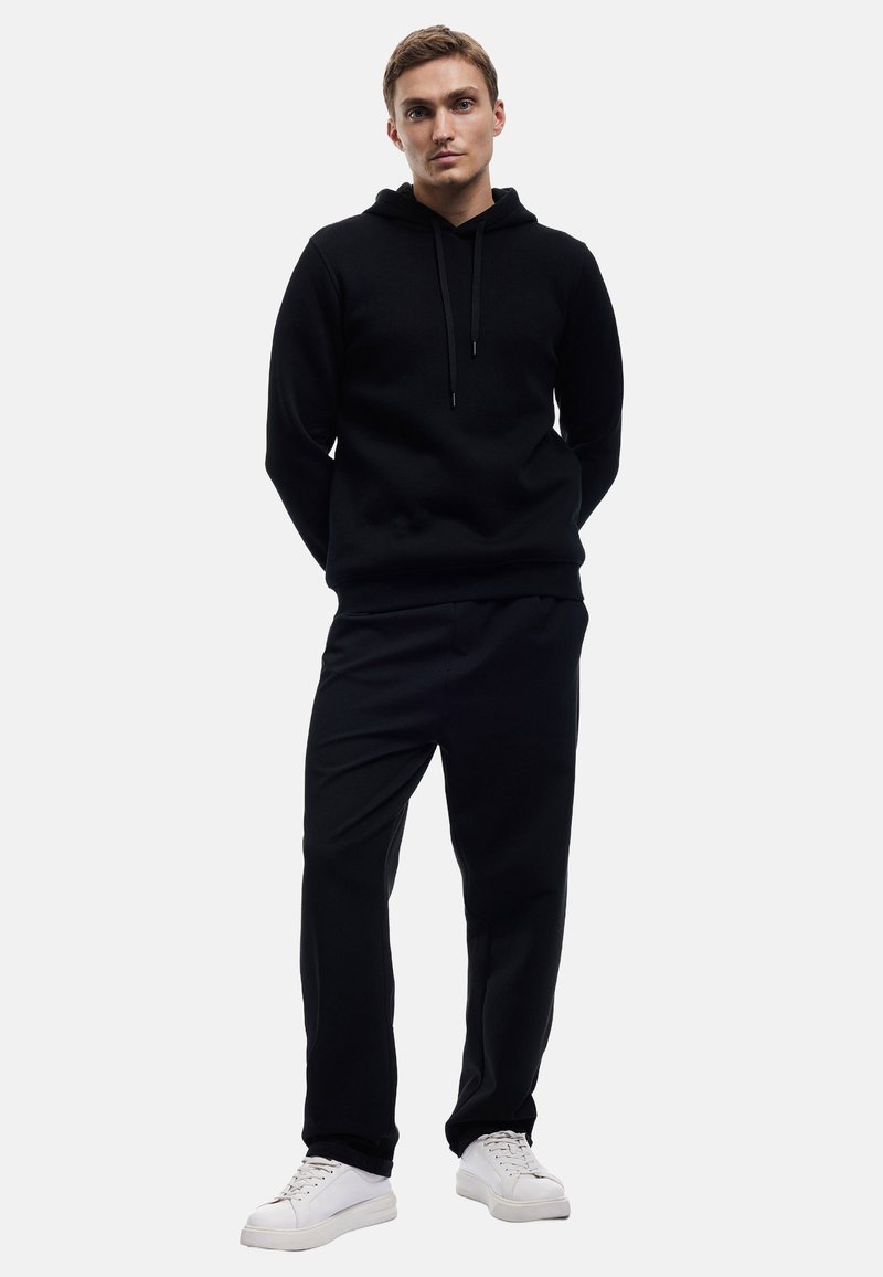 Sort hættetrøje med snørre, parret med sorte bukser. Begge dele er lavet af blødt stof. Modellen har hvide sneakers på og står foran en ensfarvet baggrund.
